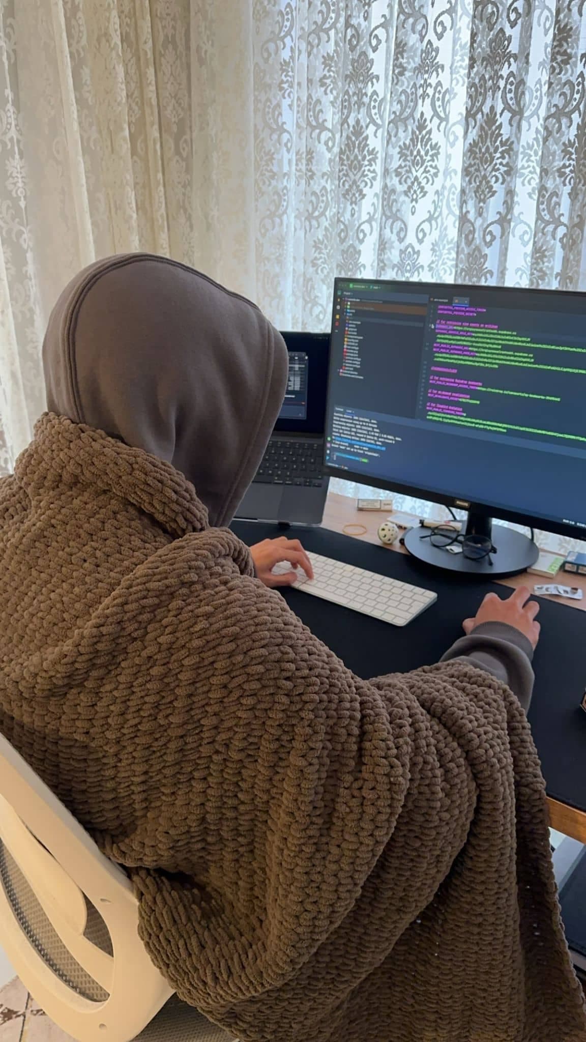 A man enveloped in a cozy blanket is focused on coding on his computer, with elegant patterned curtains in the background allowing natural light to illuminate the room.