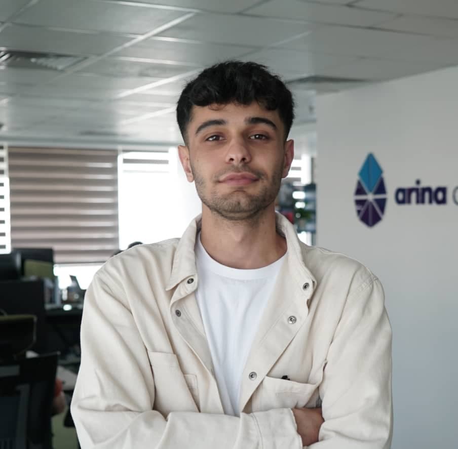 A confident young man with a casual hairstyle, wearing a white t-shirt and a light jacket, stands with crossed arms in a modern office environment, with the company logo visible in the background.