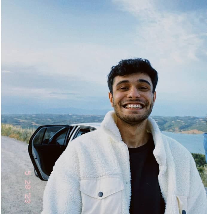 A cheerful young man wearing a cozy sherpa jacket smiles broadly, with a serene landscape and an open car door in the background, suggesting the start of an adventure.